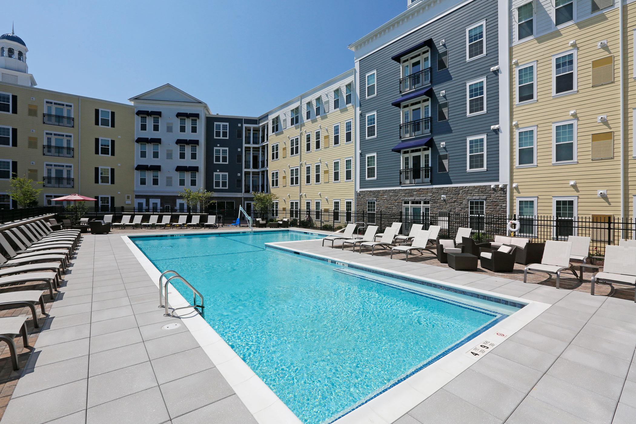 An outside view of an L shaped pool surrounded by pool chairs, and apartments with windows.