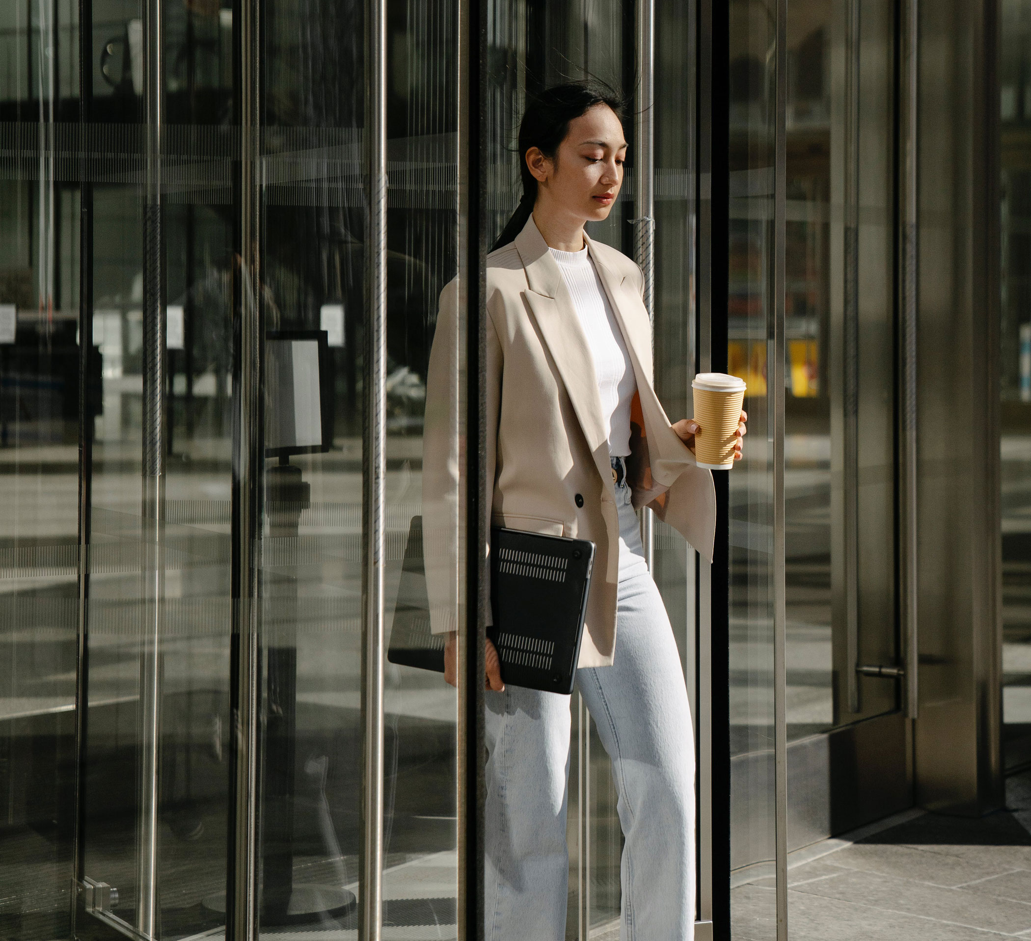 A woman holding a laptop in one hand and to-go coffee mug walking out a glass door.