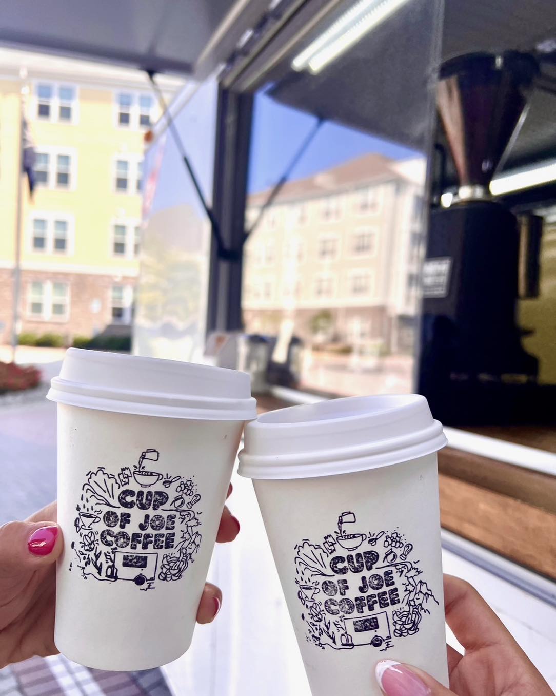Two manicured hands cheers their to-go coffee cups, showing the "Cup of Joe Coffee" art.