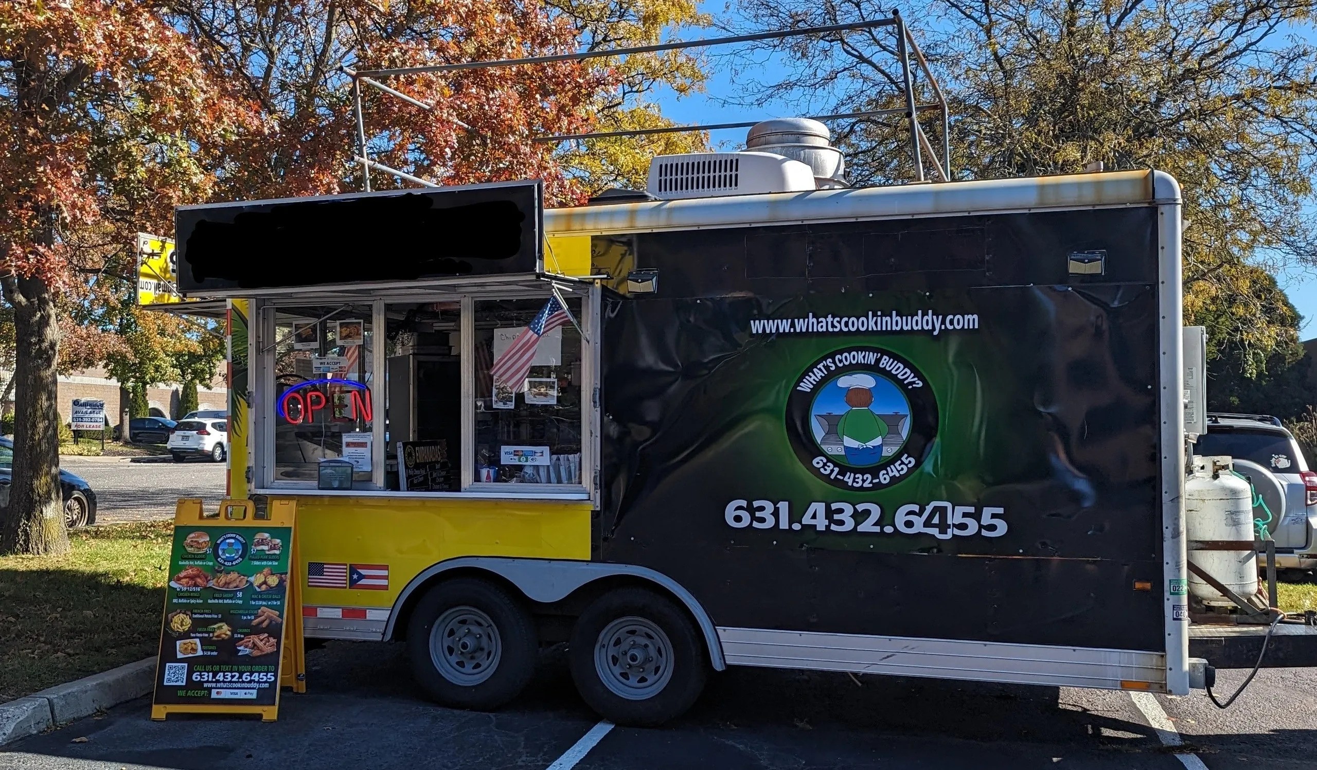 A yellow and green "What's Cooking Buddy" Food Truck in a parking lot with a food menu propped up.